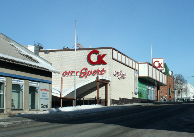 Ein Sportgeschäft in einer Stadt, das an einer Straßenecke liegt und von Gebäuden, Straßenlaternen, Bäumen und Schnee umgeben ist, mit dem Himmel im Hintergrund.