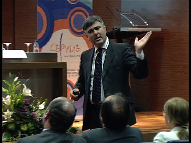 Ein Mann in einem schwarzen Blazer steht an einem Podium mit Mikrofonen, spricht in eines hinein und hat ein Blumentopf oder einen Blumenstrauß in der Nähe. Vor ihm sitzen zwei Männer und eine Frau auf Stühlen. Hinter ihm ist eine braune Wand und ein buntes Schild mit Text zu sehen, was auf eine Tagungsraum-Atmosphäre hindeutet.