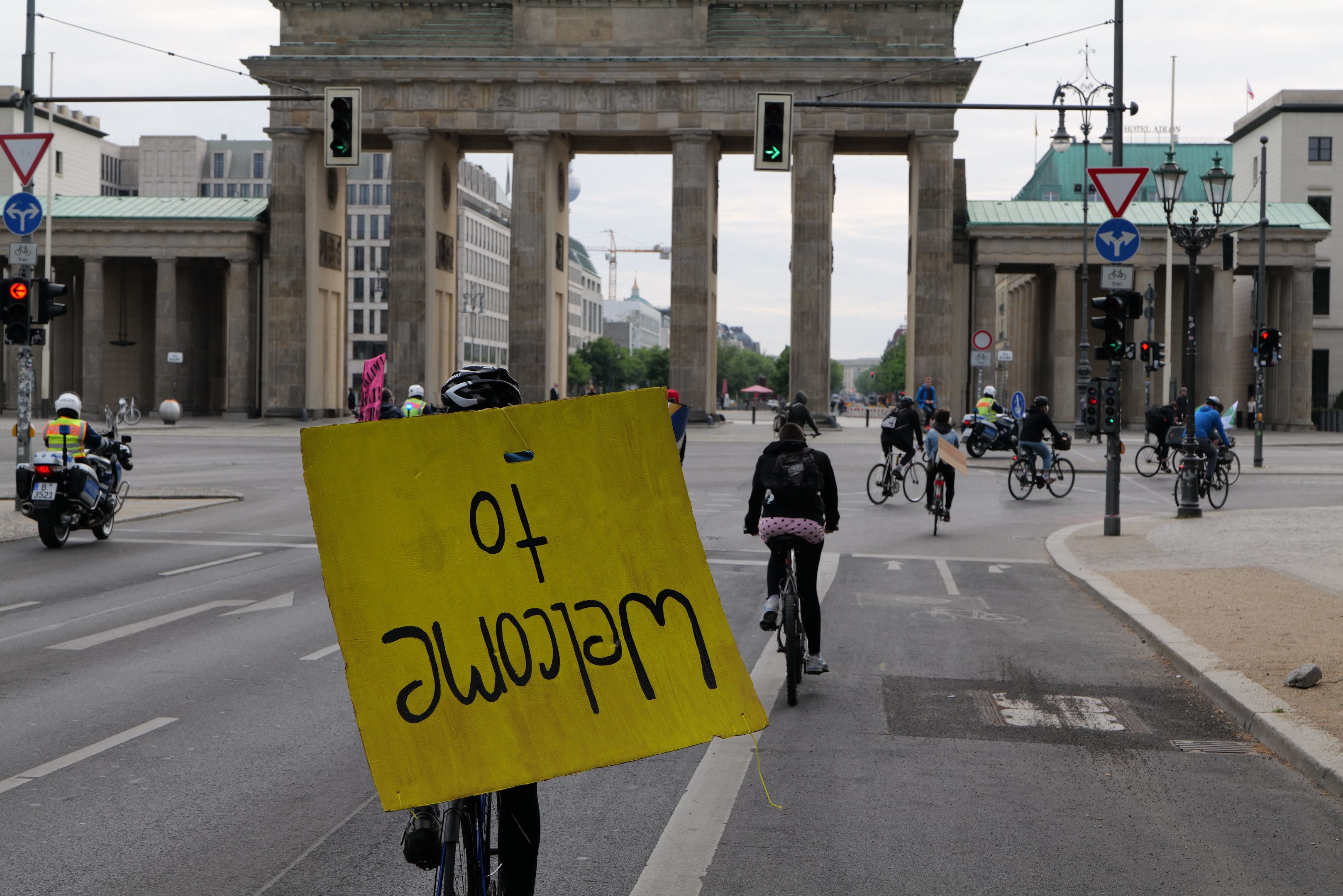 Eine Gruppe von Radfahrern fährt mit Helmen die Straße vor dem Brandenburger Tor in Berlin, Deutschland, entlang, wobei eine Person ein gelbes Schild hält, Lichtmasten, Verkehrszeichen, Gebäude, Bäume und einen klaren blauen Himmel im Hintergrund.