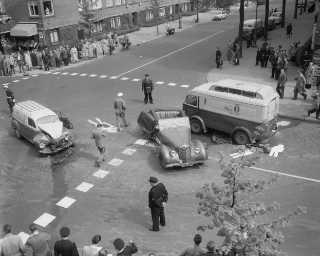 Schwarz-weißes Bild eines Autounfalls an einer Straßenkreuzung mit mehreren Fahrzeugen, darunter ein Van, und einer Gruppe von Menschen, die daneben stehen, mit Gebäuden, Bäumen, Pfählen und Texttafeln im Hintergrund.