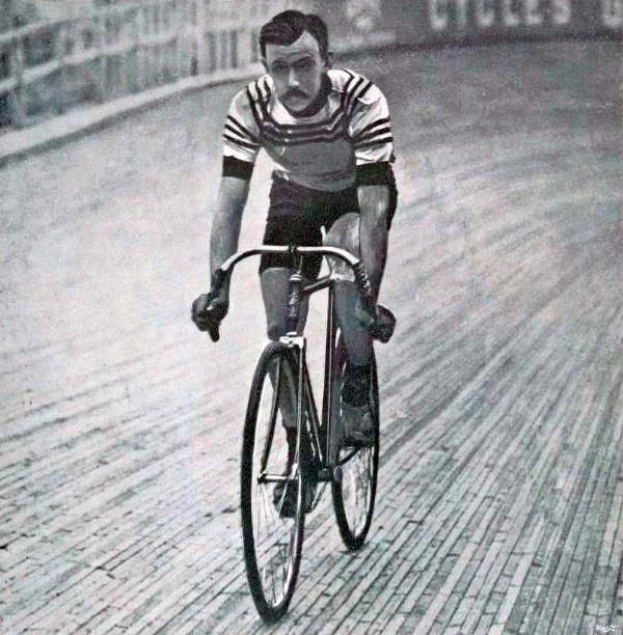 Altes Schwarz-Weiß-Foto eines Mannes, der auf einer Bahn ein Fahrrad fährt, mit einem Zaun und einer Tafel im Hintergrund.