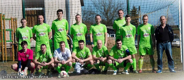Eine Gruppe von Männern in grünen T-Shirts steht auf einem Fußballfeld in der Nähe eines Torpfostens, mit einem Ball vor ihnen und verschiedenen Hintergrundelementen wie Bäume, Gebäude und Fahrzeuge.