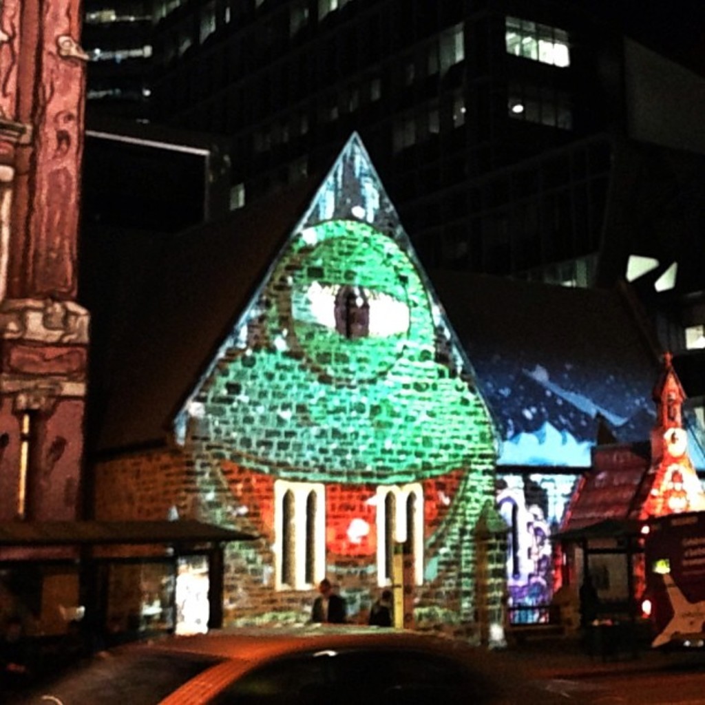 Ein zentrales Haus mit einer Person daneben, flankiert von Häusern auf beiden Seiten, ein Gebäude mit Lichtern im Hintergrund und ein Auto unten.