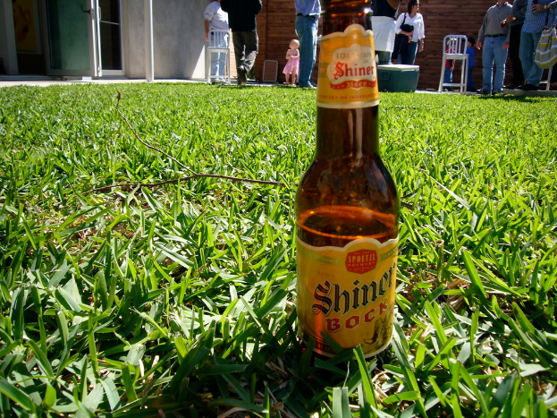 Eine Bierflasche mit der Aufschrift "Shiner Bock" steht auf Gras, mit Menschen, Stühlen und einem Haus im Hintergrund.