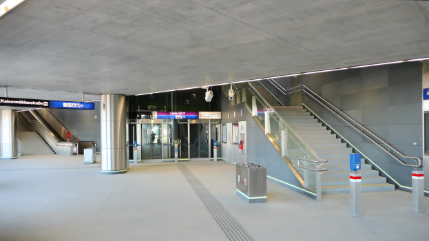 Leerer Bahnhof mit Treppe, Geländern, Mülltonnen, Informationsschildern, Deckenlampen und Glastüren im Hintergrund.