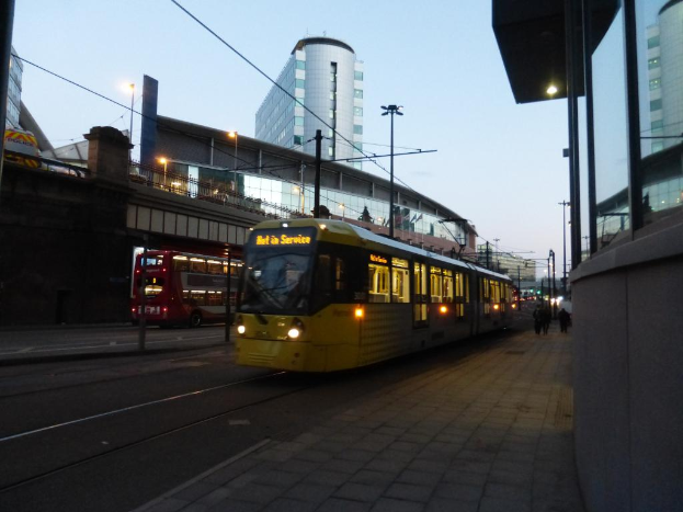 Ein Bus fährt auf einer Straße mit Gebäuden auf beiden Seiten, jeweils mit Glasfenstern, und einer Brücke auf der linken Seite. Es gibt auch Laternenpfähle entlang der Straße.