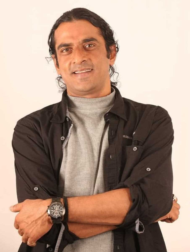 Ein Mann in einem schwarzen und aschfarbenen Kleid mit überschlagenen Armen, der eine Uhr tr├Ągt, vor einem wei├čen Hintergrund steht und nachdenklich wirkt.