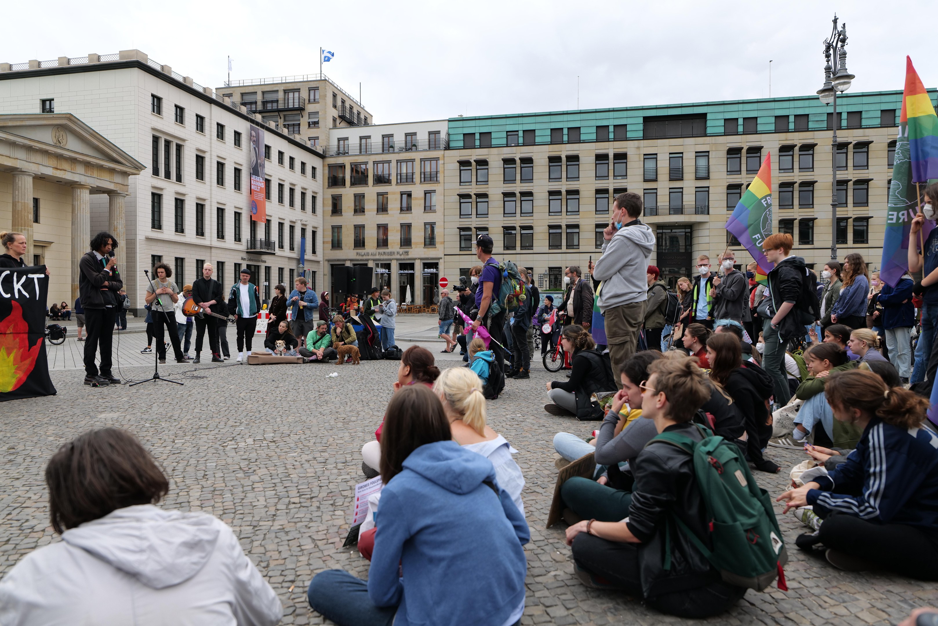 Eine Gruppe von Menschen sitzt auf dem Boden vor einer Menge, die Fahnen und Plakate hält, während einer Anti-Schwulen-Demonstration in Berlin. Im Hintergrund sind eine Statue, ein Mikrofonständer und Gebäude zu sehen.