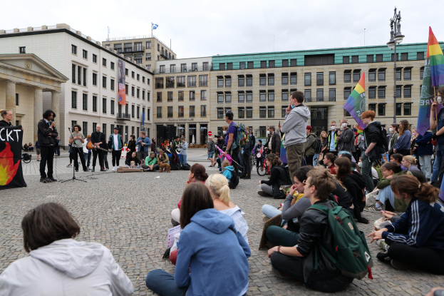 Eine Gruppe von Menschen sitzt auf dem Boden vor einer Menge, die Fahnen und Plakate hält, während einer Anti-Schwulen-Demonstration in Berlin. Im Hintergrund sind eine Statue, ein Mikrofonständer und Gebäude zu sehen.