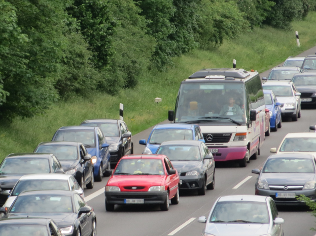 Verkehrsstau auf einer Autobahn mit Autos und einem Lieferwagen, Menschen in den Fahrzeugen, Bäume und Gras im Hintergrund.