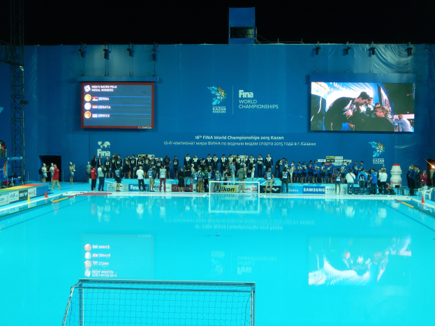 Gruppe von Menschen um ein Hallenbad mit Netz am Boden herumstehend mit einer Wand im Hintergrund, die den Text "FINA World Championships 2015 Kazan" und Logos, Träger und Lampen anzeigt.
