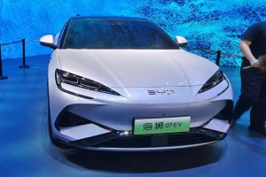 Ein weißes Auto auf einer Autauschau mit einer Person rechts daneben, die ein Objekt hält, ein Bildschirm im Hintergrund und Absperrpoller mit Seilen links.