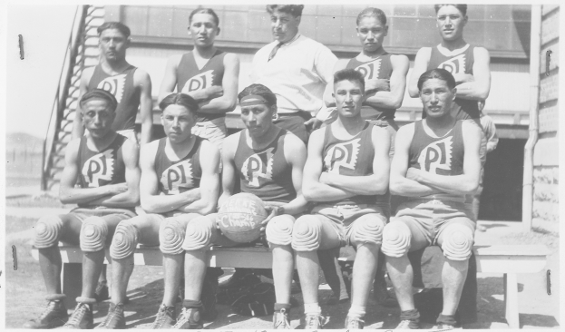 Schwarzes und weißes Foto einer Basketballmannschaft, die auf einer Bank sitzt, mit einem Mann in der Mitte, der einen Ball hält, Treppen und eine Wand im Hintergrund und Text unten.