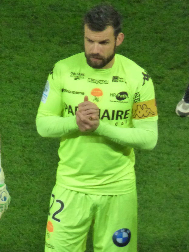 Ein Torwart in einem gelben Trikot klatscht auf einem Fußballfeld, trägt ein grünes Hemd und kurze Hosen und hat einen Teil eines anderen Beins im Bild.