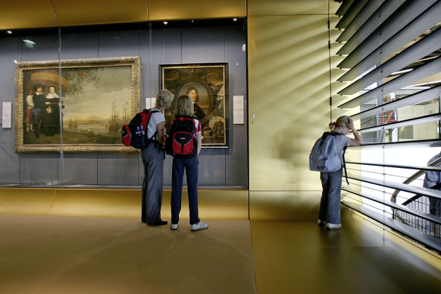 Drei Personen in einem Museum, das Gemälde an der Wand betrachtet, mit einem Geländer auf der rechten Seite und warmem Deckenlicht.
