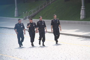 Drei Polizisten in Uniform und Mützen, die eine Straße mit Pfählen, Geländern, Treppen und Gras im Hintergrund entlanggehen.