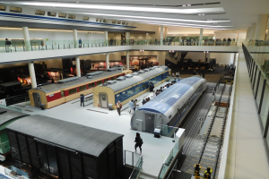 Ein geräumiger Museumsraum mit zahlreichen Zügen in verschiedenen Größen und Farben, bei dem Besucher die Ausstellungsstücke, Geländer, Säulen und Deckenleuchten betrachten.