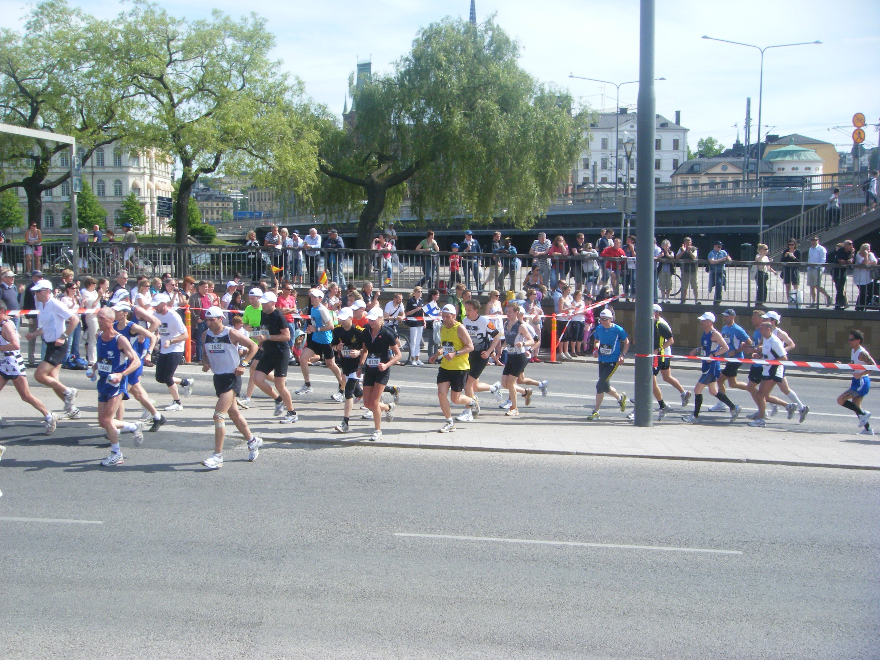Gruppe von Läufern bei einem Marathon auf einer Straße mit Metallzaun, Tor, Band, Zuschauern, Absperrungen, Pfosten, Straßenschildern, Brücke, Gebäuden, Bäumen und bewölktem Himmel.