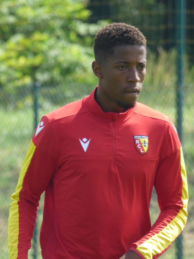 Ein junger Mann in einem roten und gelben Trikot steht auf einem Fußballfeld, umgeben von einem Zaun und Bäumen, und sieht entschlossen aus.