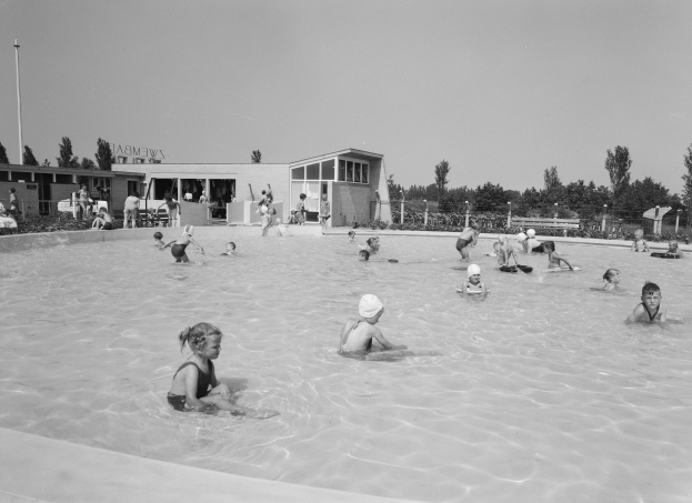 Schwarzes und weißes Foto von Kindern, die in einem Swimmingpool spielen, umgeben von Gebäuden, Bäumen, Pfählen, Pflanzen und einem Zaun unter einem klaren Himmel.