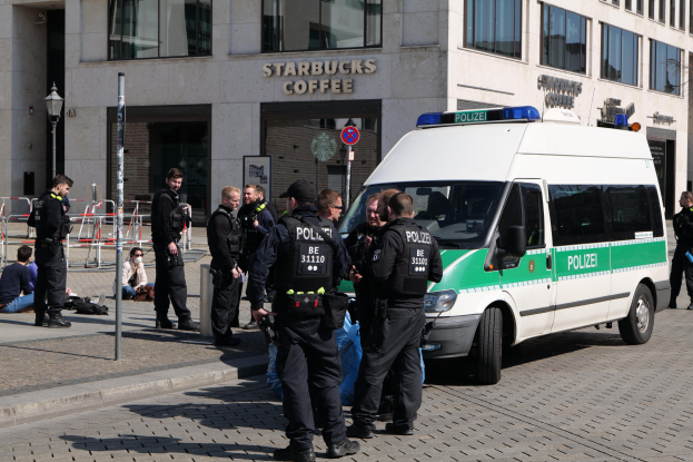 Eine Gruppe von Polizeibeamten vor einem Starbucks-Café mit einem Van rechts und Menschen links, vor einem Hintergrund mit Fenstern, einem Schild, einem Laternenmast und einem Zaun.