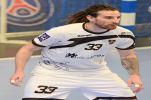 Ein Mann mit langen Haaren und Arm-Tattoos spielt Handball in einem weißen T-Shirt und Shorts, mit einem Netz und einer Wand im Hintergrund.