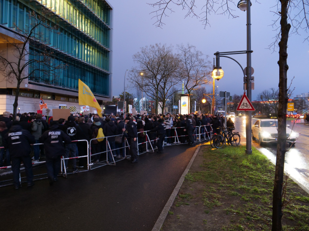 Eine große Gruppe von Menschen protestiert vor einem Gebäude in Berlin, hält Schilder und steht hinter Barrikaden, mit Fahrrädern, Laternen, Schildern, Bäumen und Gras im Hintergrund.