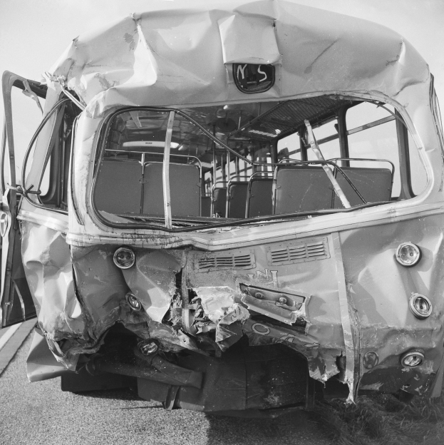 Ein Schwarz-Weiß-Foto eines schwer beschädigten Busses am Straßenrand, dessen Vorderseite eingedrückt und Fenster zerbrochen sind, vor einem sichtbaren Himmel.