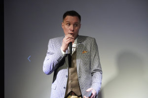 Ein Mann in einem maßgeschneiderten Anzug steht selbstbewusst auf einer Bühne, hält ein Mikrofon und hat einen entschlossenen Gesichtsausdruck sowie ordentlich gekämmtes Haar.