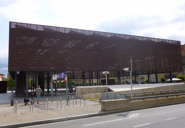 Großes modernes Gebäude mit zahlreichen Fenstern, das als neuer Eingang zur Stadt dient, umgeben von Straßeninfrastruktur, Fahrzeugen, Fußgängern, Bäumen und einem bewölkten Himmel.