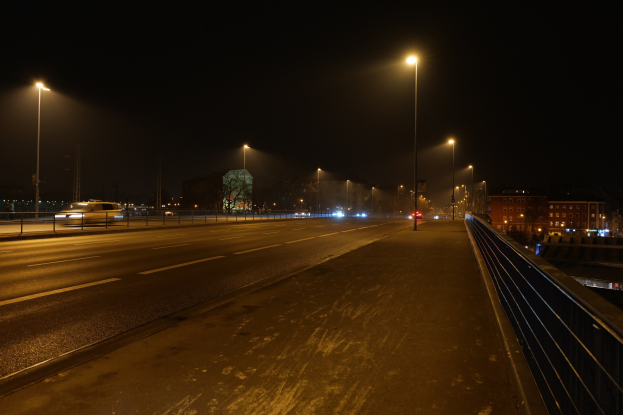 Langzeitbelichtungsfotografie einer nächtlichen Stadtstraße, die sich bewegende Fahrzeuge, Geländer, Laternenpfähle, Gebäude, Bäume und einen dunklen Himmel zeigt.
