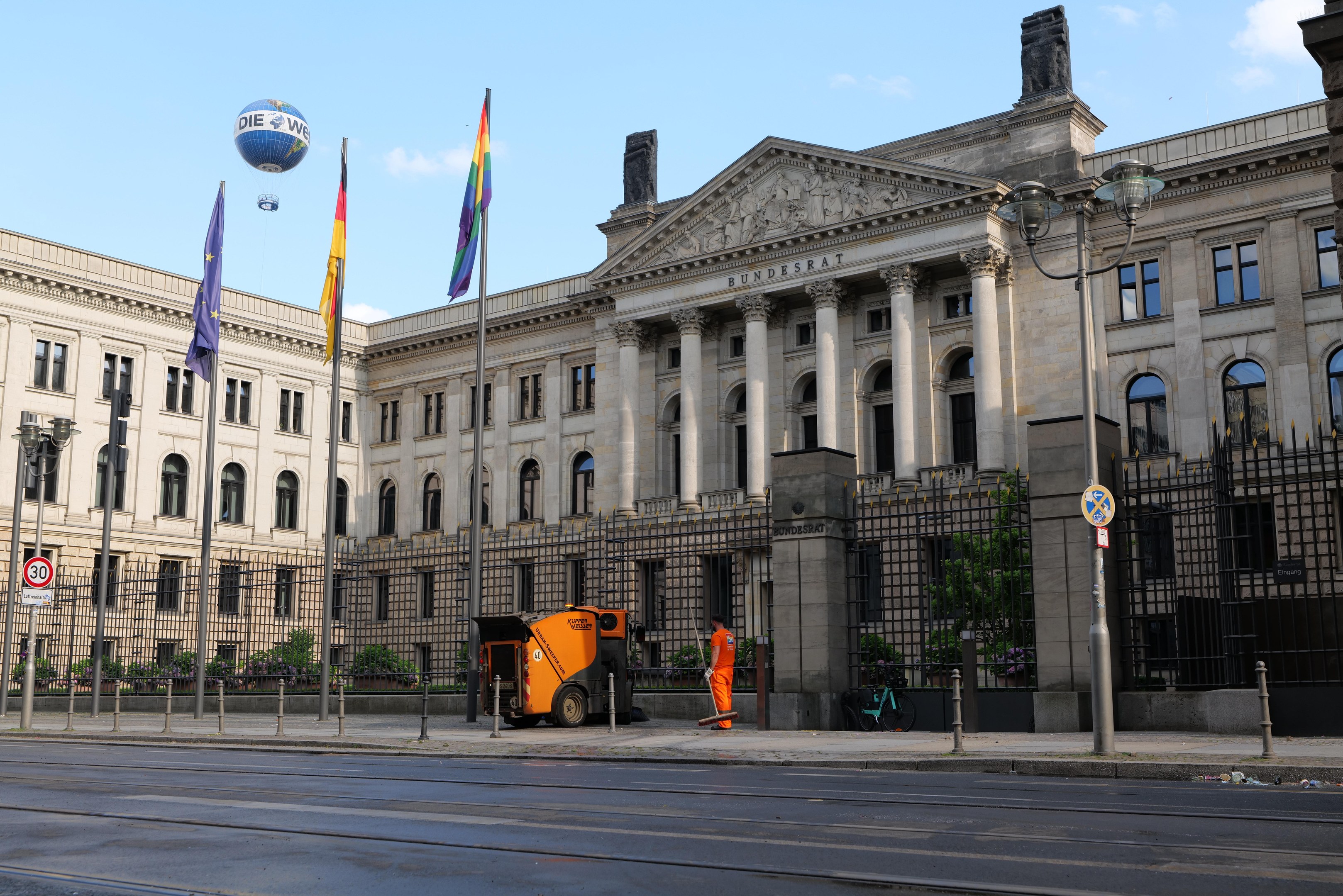 Großes Regierungsgebäude mit vielen Fenstern, umgeben von Straßeninfrastruktur und Fußgängern, mit Flaggen und Bäumen davor, unter einem bewölkten Himmel.