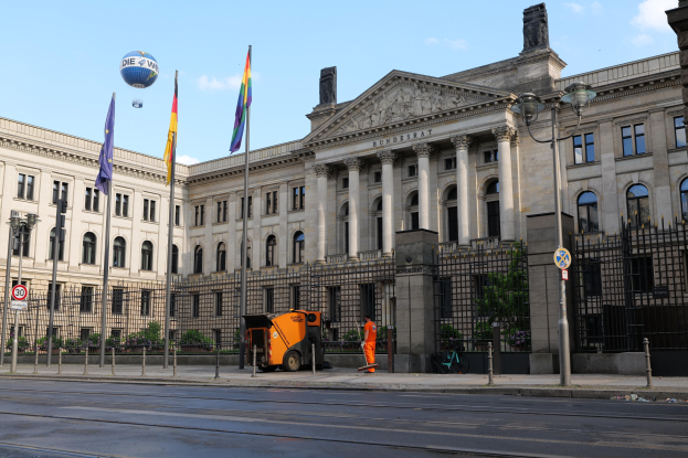 Großes Regierungsgebäude mit vielen Fenstern, umgeben von Straßeninfrastruktur und Fußgängern, mit Flaggen und Bäumen davor, unter einem bewölkten Himmel.