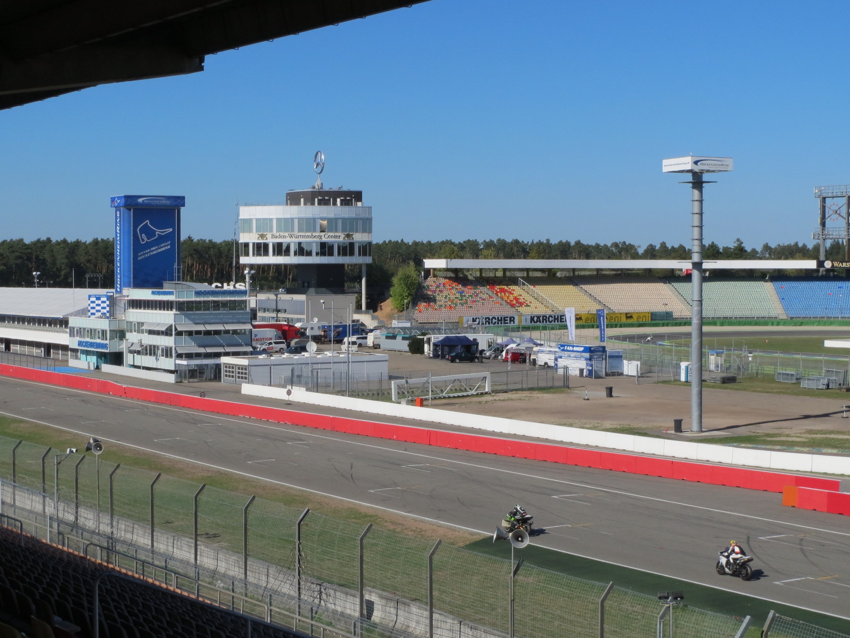 Eine Gruppe von Menschen auf Motorrädern auf einer Rennstrecke umgeben von einem Metallzaun, Fahrzeugen, einem Gebäude, einem Schild, Pfosten, einem Stadion, Lichtern, Bäumen und einem bewölkten Himmel.