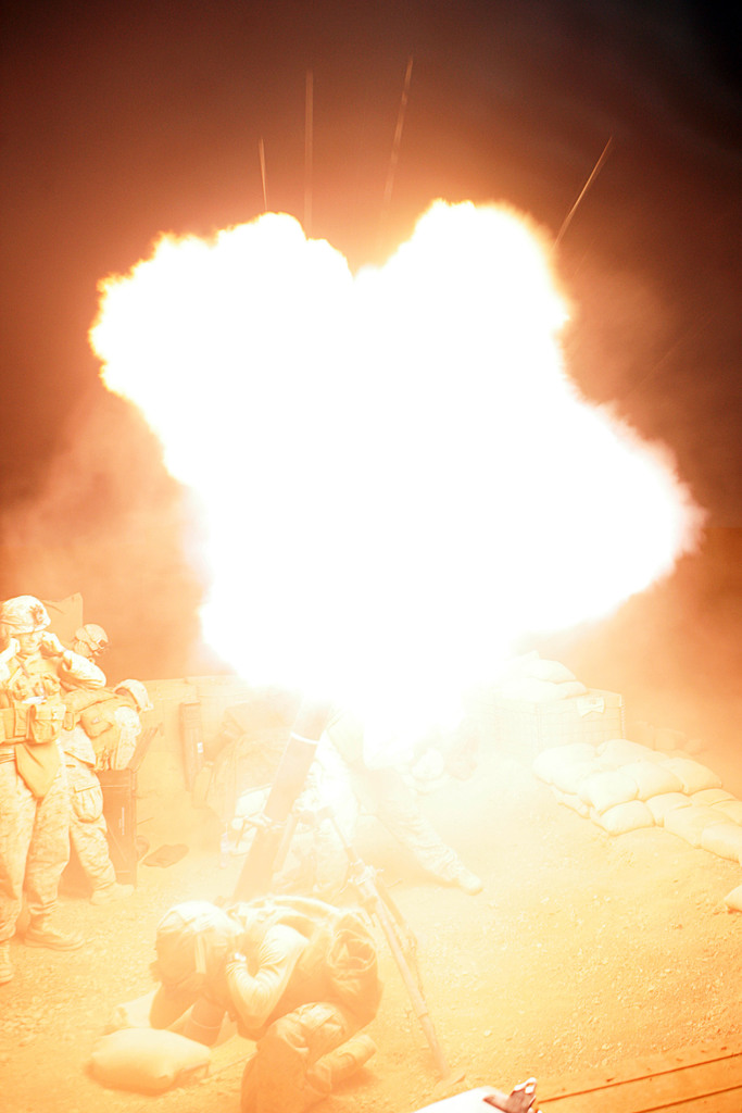 Mehrere Personen in Uniformen und Helmen befinden sich auf einem Gelände mit einigen Taschen, die herumliegen, und einem Feuer in der Mitte der Szene.