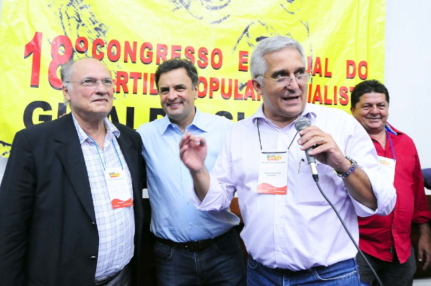 Ein Mann spricht in ein Mikrofon vor einem gelben Vorhang mit der Aufschrift "18-Grad-Kongress", während andere Männer im Hintergrund stehen.
