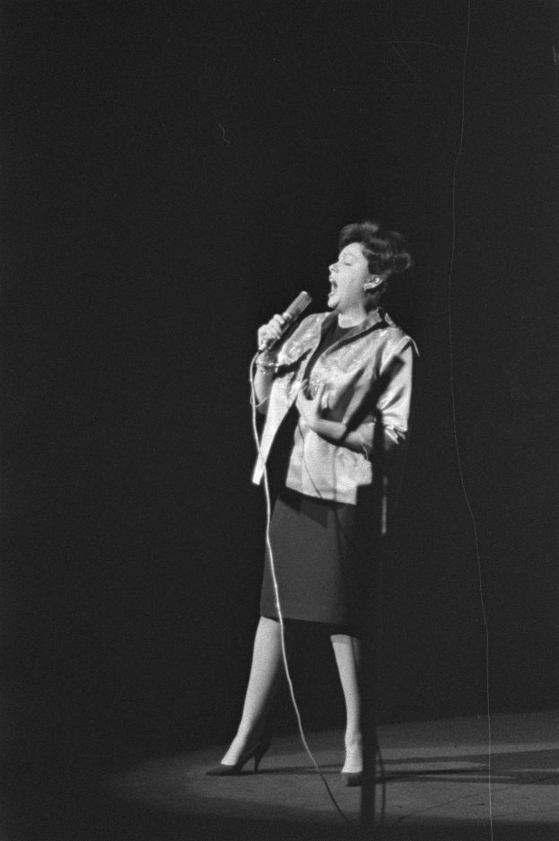 Eine Frau in einem schwarz-weißen Outfit aus den 1950er-Jahren singt fröhlich auf einer von einem Scheinwerfer beleuchteten Bühne in ein Mikrofon, ihr Haar ist im klassischen 50er-Jahre-Stil frisiert.