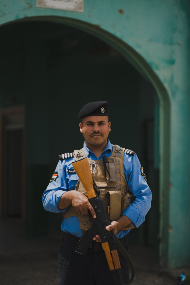 Polizeibeamter in Uniform mit Gewehr, vor einem Gebäude mit einem Torbogen und einer Tafel an der Wand stehend.