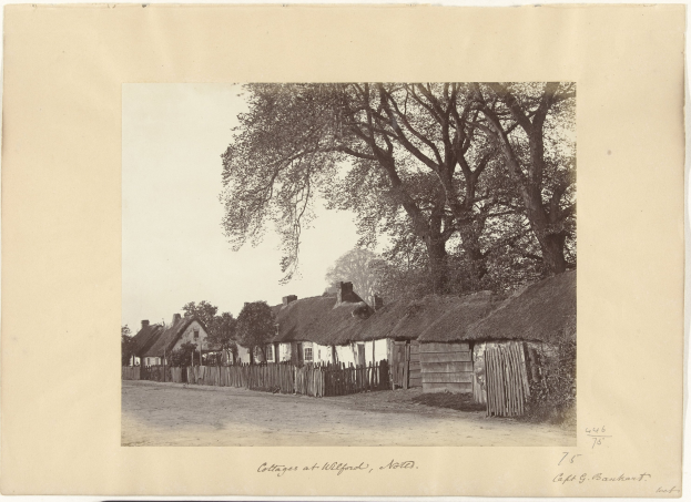 Schwarz-weiß-Foto eines Dorfs mit Reetdächern, einer Straße, einem Zaun, Bäumen und einem Himmel im Hintergrund, mit Text unten.