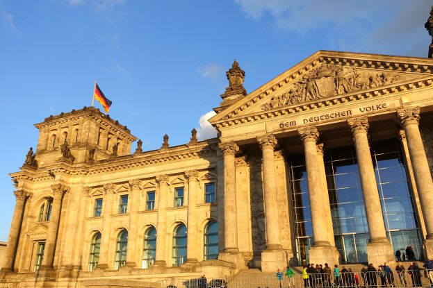 Das Reichstagsgebäude in Berlin, Deutschland, ein großes Gebäude mit Fenstern, Säulen und einer Flagge oben drauf, mit einem Geländer davor und ein paar Menschen drumherum, unter einem bewölkten Himmel.