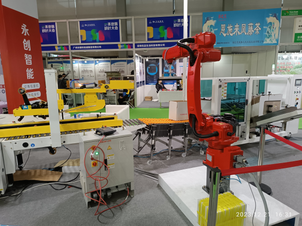 Gruppe von Robotern auf einem Tisch in einer Fabrikumgebung umgeben von Maschinen, Kabeln, textbedruckten Platinen und beleuchteten Komponenten, mit Menschen im Hintergrund.