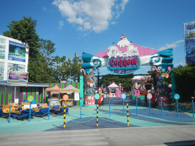 Kirmesfahrgeschäft am Parkeingang mit Straße, eingezäuntem Bereich, Buden, Bannern, Bäumen und bewölktem Himmel.