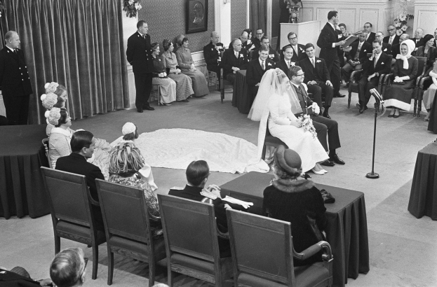 Schwarzes und weißes Foto einer Hochzeitszeremonie mit Menschen, die sitzen und stehen, einem Mann mit Mikrofon, einer Frau mit einem Strauß, einem Podium mit Mikrofon, Blumenschmuck, einem Vorhang, einem gerahmten Foto an der Wand und einer Tür im Hintergrund.