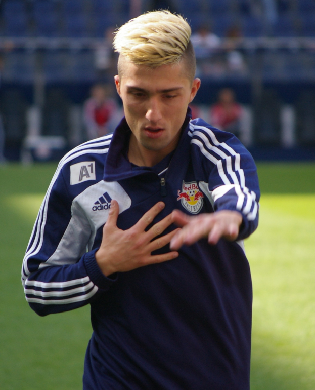 Ein junger Mann in einem blauen T-Shirt steht auf einem Fußballfeld und streckt die Arme aus, wobei er entschlossen als Flügelspieler mit einem unscharfen Hintergrund erscheint.