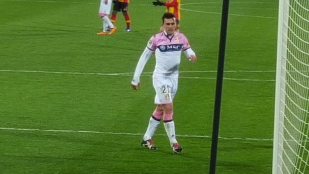 Ein Fußballspieler steht auf einem Feld neben einem Tor mit Netz auf der rechten Seite, während andere Spieler im Hintergrund sichtbar sind, was ein laufendes Spiel andeutet.