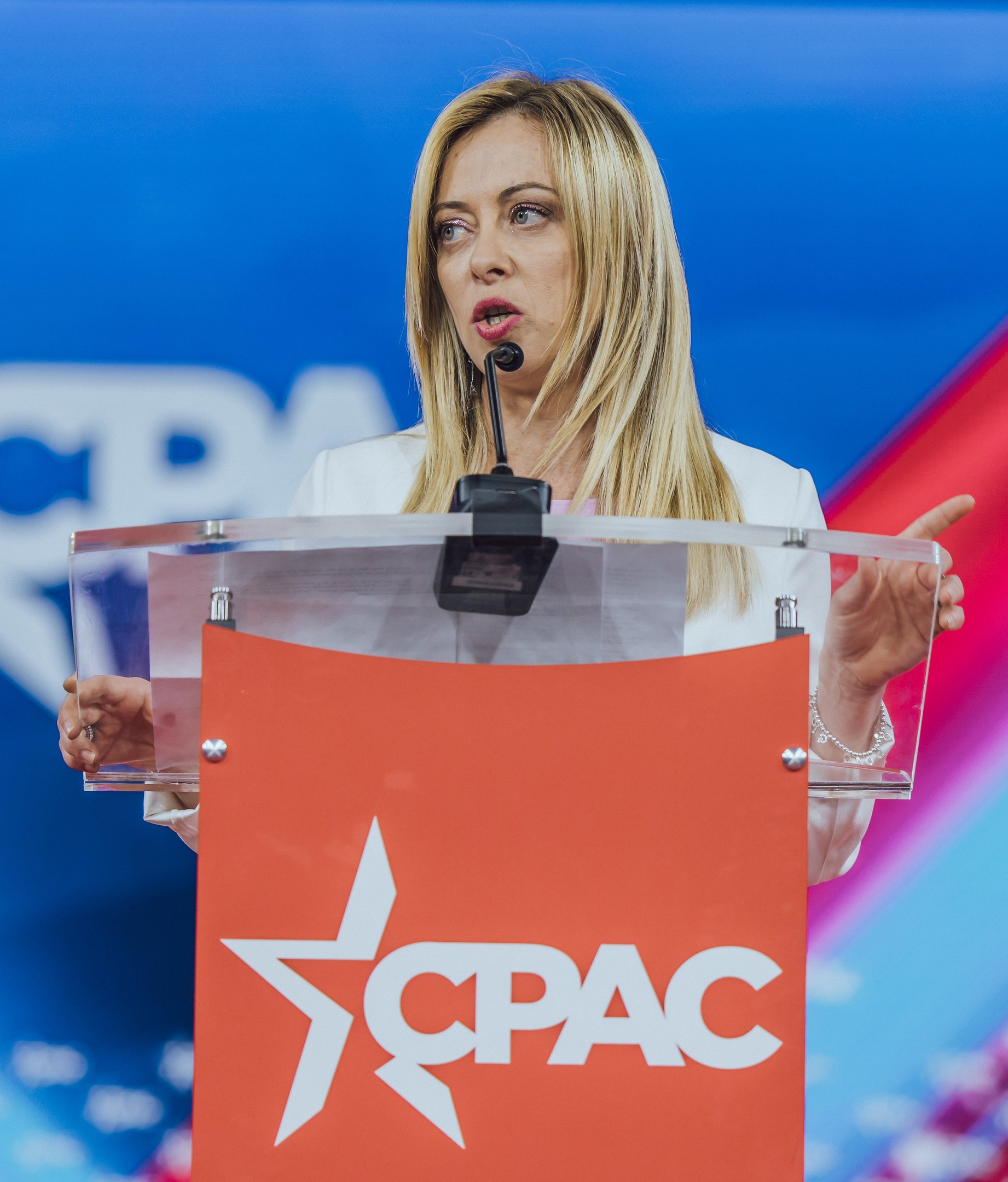 Frau in weißem Kleid, die an einem Rednerpult mit einem Mikrofon spricht, unscharfer Hintergrund.