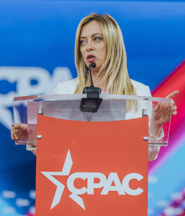 Frau in weißem Kleid, die an einem Rednerpult mit einem Mikrofon spricht, unscharfer Hintergrund.