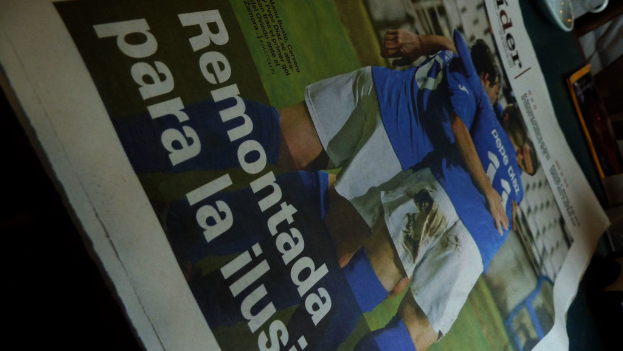 Eine Zeitung auf einem Tisch zeigt drei M├Ąnner in blauen Hemden, eventuell beim Fu├čballspielen, mit Text am unteren Rand des Bildes.