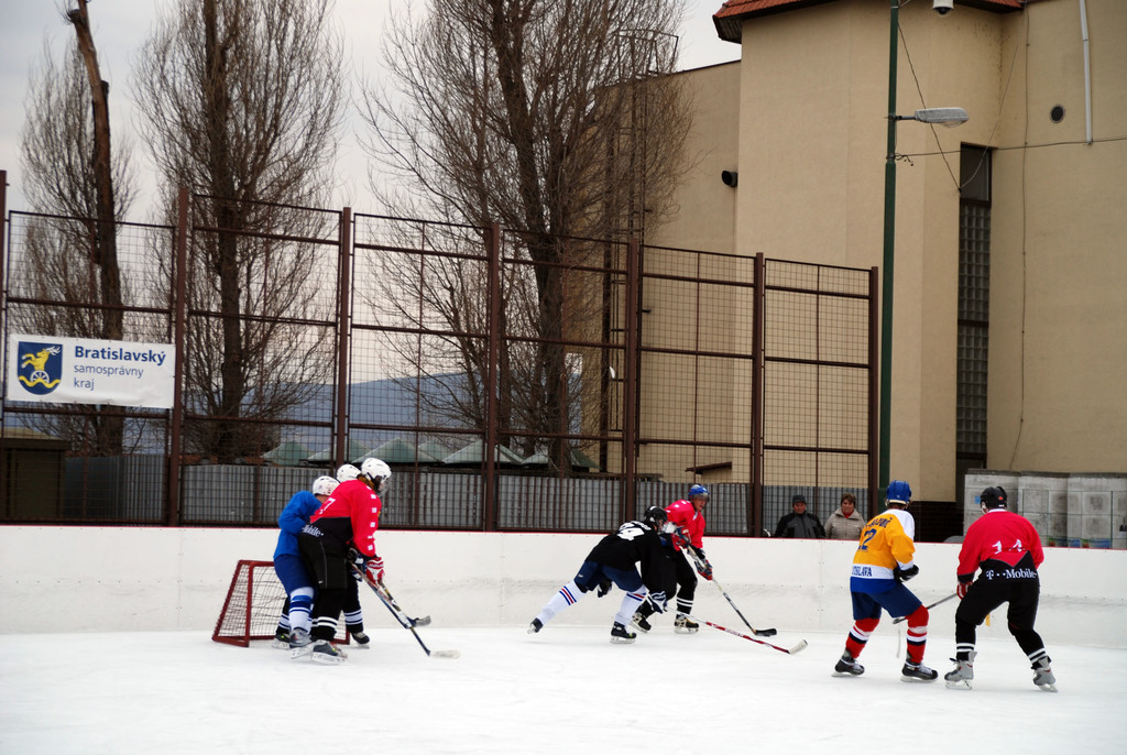 Personen spielen Eishockey auf einem Eisplatz mit Gebäuden, Bäumen, einer Straßenlaterne, einem Namensschild und Zäunen im Hintergrund unter einem Himmel.