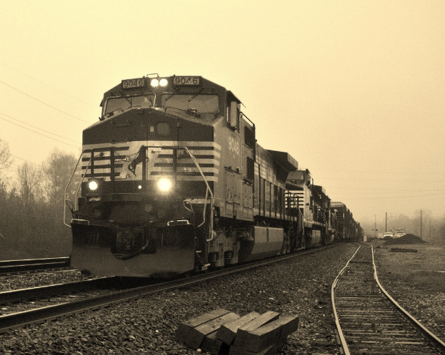 Ein Zug auf Eisenbahnschienen mit Autos, Strommasten, Bäumen und einem Himmel im Hintergrund.